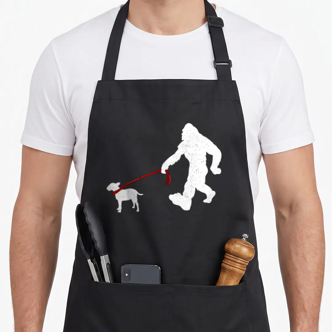 Bigfoot dog dad apron - White Bigfoot silhouette walking a small dog on a red leash with distressed texture — shown on a black cotton canvas apron against a white background