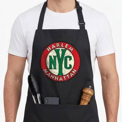 Harlem nyc apron - Distressed circular emblem with HARLEM NYC MANHATTAN in red green cream black lettering — shown on a black cotton canvas apron against a white background