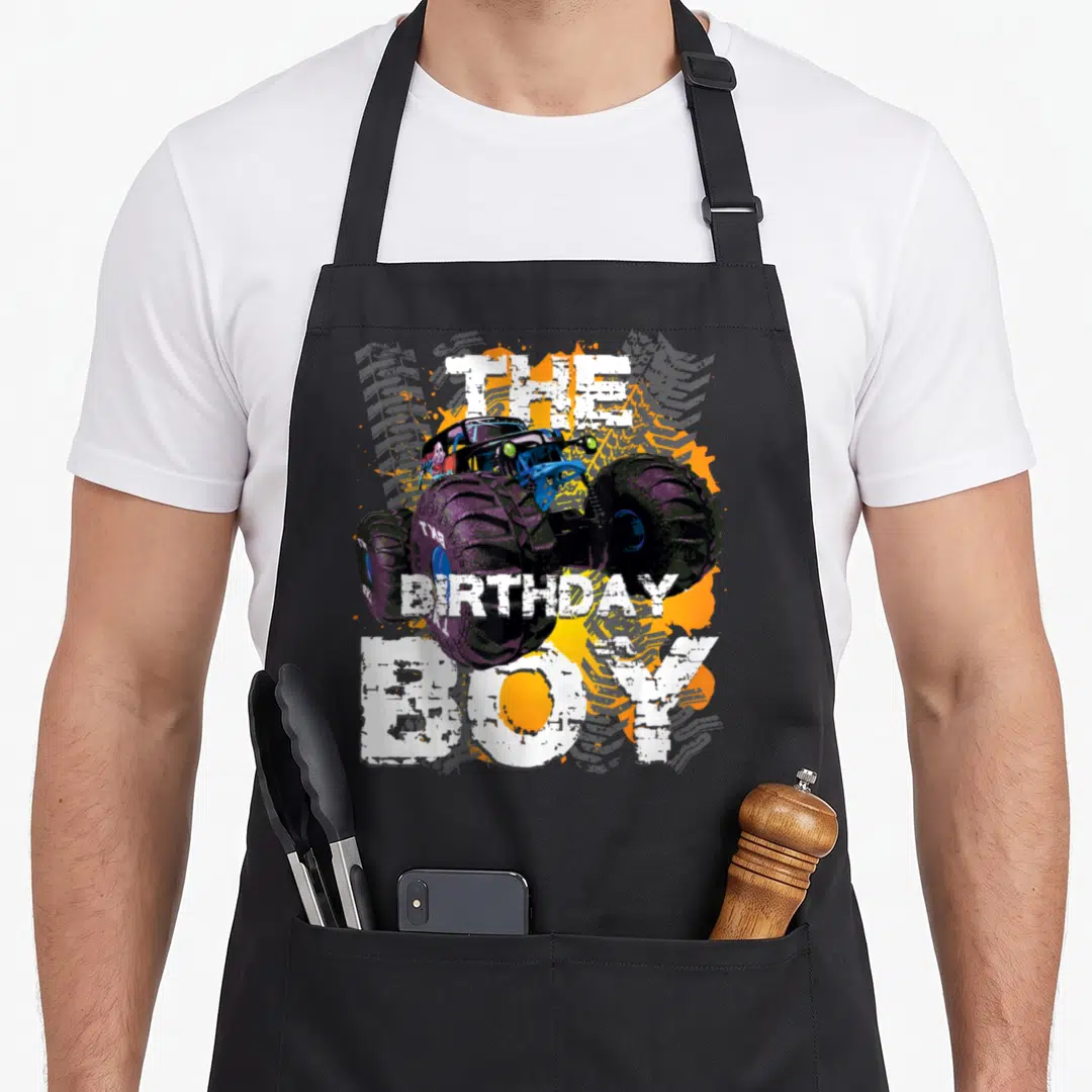 Family truck apron - Monster truck with purple tires and white The Birthday Boy text over orange splatters — shown on a black cotton canvas apron against a white background