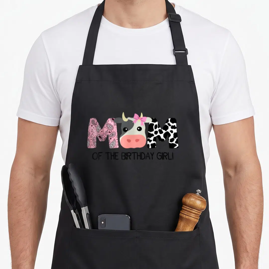 Farm cow print apron - Cartoon cow head replaces O in MOM text with pink bow and patterned letters — shown on a black cotton canvas apron against a white background