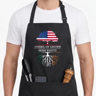 Root ireland apron - Tree with US flag leaves and Irish flag roots separating text AMERICAN GROWN WITH IRISH ROOTS — shown on a black cotton canvas apron against a white background