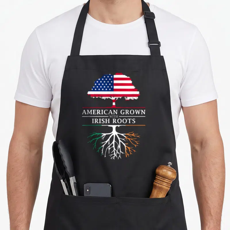 Root ireland apron - Tree with US flag leaves and Irish flag roots separating text AMERICAN GROWN WITH IRISH ROOTS — shown on a black cotton canvas apron against a white background