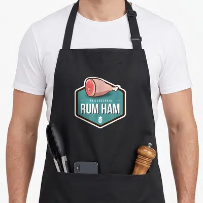 Ham funny rum apron - Cartoon ham on teal hexagon with PHILADELPHIA RUM HAM text and small pineapple icon — shown on a black cotton canvas apron against a white background