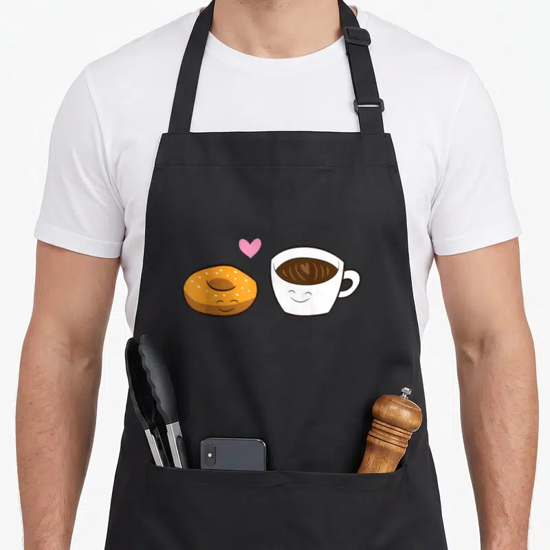 Cute bagel coffee apron - Cartoon donut and coffee cup with smiling faces and pink heart — shown on a black cotton canvas apron against a white background