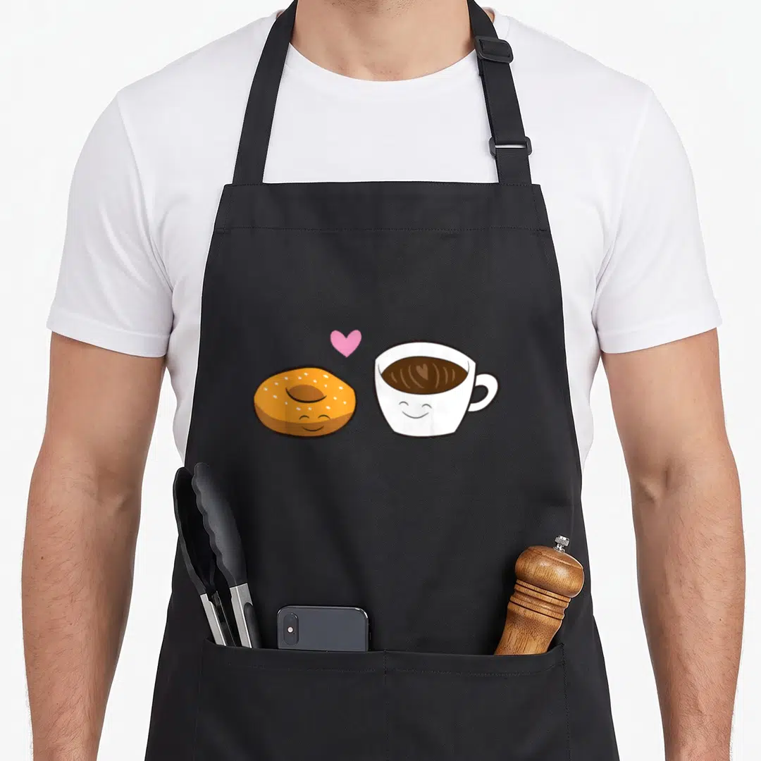 Cute bagel coffee apron - Cartoon donut and coffee cup with smiling faces and pink heart — shown on a black cotton canvas apron against a white background