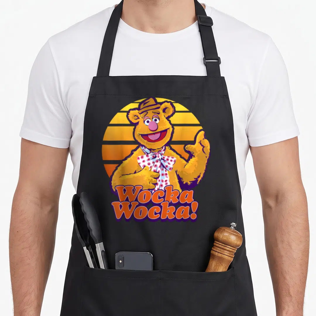 Fozzie bear apron - Cartoon bear with striped sun background and Wocka Wocka text in orange and purple — shown on a black cotton canvas apron against a white background