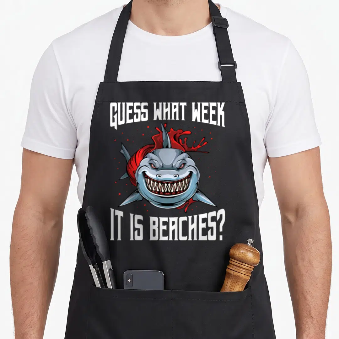Shark joke apron - Cartoon shark head with red splashes and white text Guess What Week It Is Beaches on grey background — shown on a black cotton canvas apron against a white background