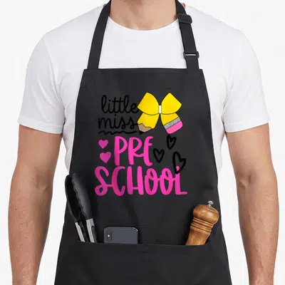 Miss preschool apron - Pencil bow graphic with little miss PRE SCHOOL text and scattered pink and black hearts — shown on a black cotton canvas apron against a white background