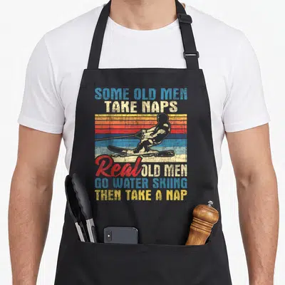 Nap grandpa apron - Retro water skier silhouette with stacked text about old men napping and skiing on colored stripes — shown on a black cotton canvas apron against a white background
