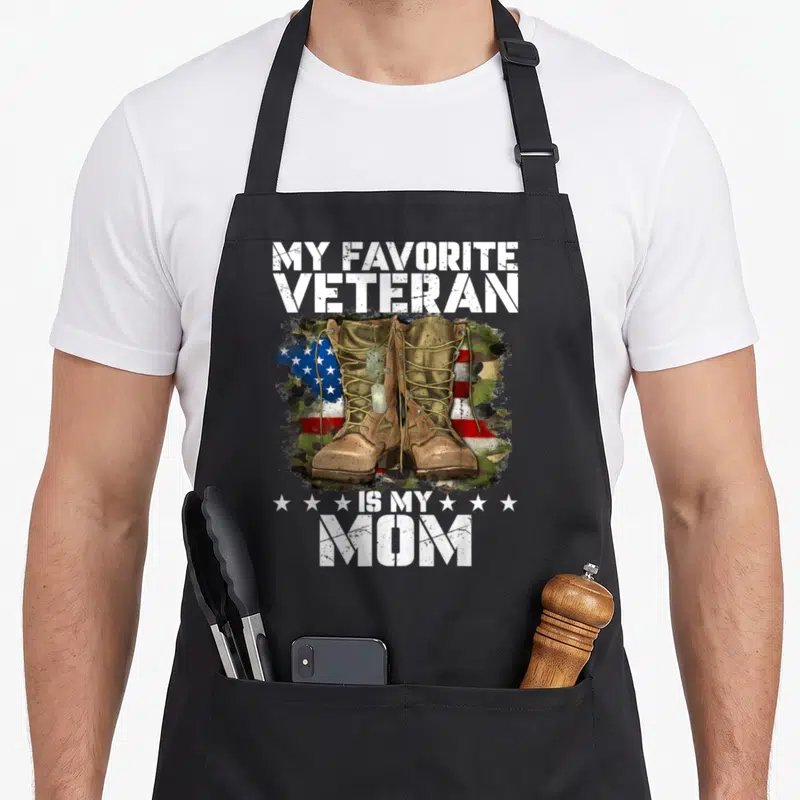 Cute veteran military apron - Tan combat boots with dog tags over distressed US flag and camouflage with white text — shown on a black cotton canvas apron against a white background