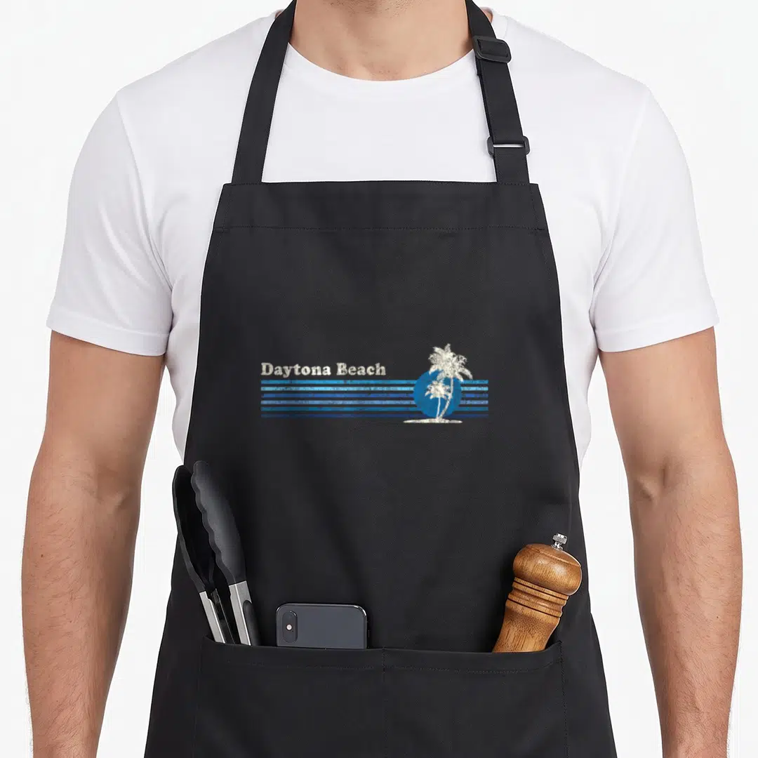 Daytona tree apron - Distressed Daytona Beach text on blue stripes with white palm tree silhouette — shown on a black cotton canvas apron against a white background