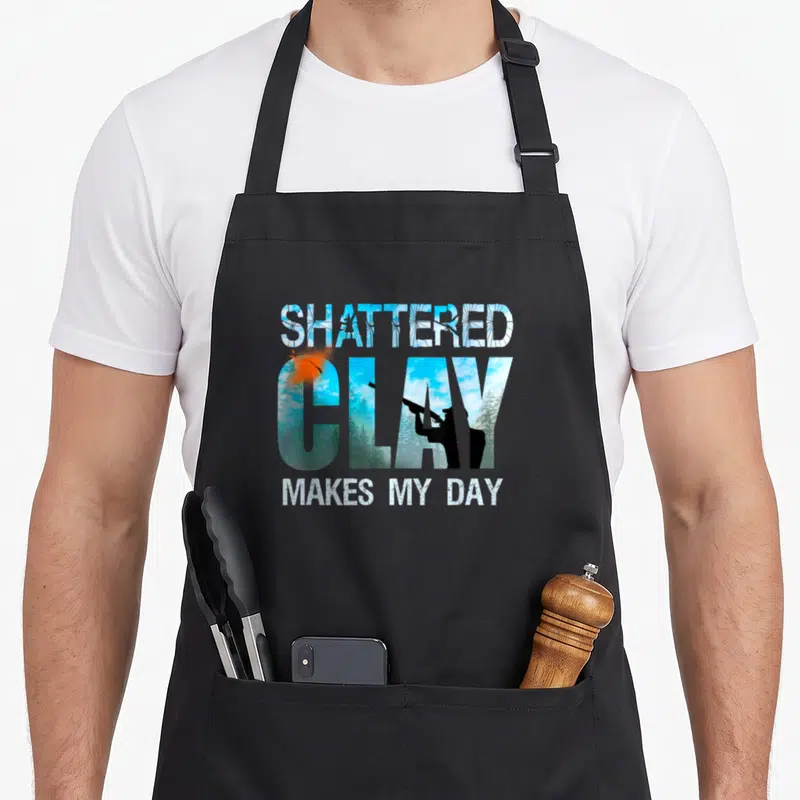 Shooting clay apron - SHATTERED CLAY MAKES MY DAY text with blue sky landscape inside letters and orange breaking clay — shown on a black cotton canvas apron against a white background