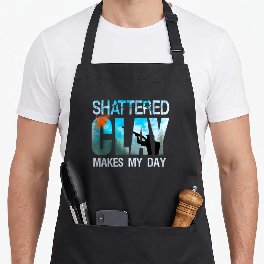 Shooting clay apron - SHATTERED CLAY MAKES MY DAY text with blue sky landscape inside letters and orange breaking clay — shown on a black cotton canvas apron against a white background