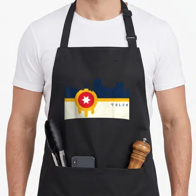 Flag tulsa apron - Dark blue Tulsa skyline over white-yellow band, red-yellow circle with white star, blue TULSA text — shown on a black cotton canvas apron against a white background