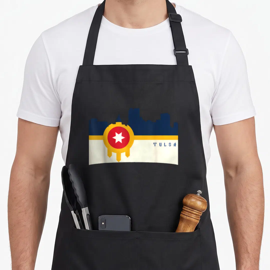 Flag tulsa apron - Dark blue Tulsa skyline over white-yellow band, red-yellow circle with white star, blue TULSA text — shown on a black cotton canvas apron against a white background