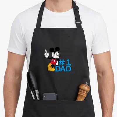 Father day apron for chefs - Mickey Mouse pointing up beside blue #1 DAD text on black background — shown on a black cotton canvas apron against a white background