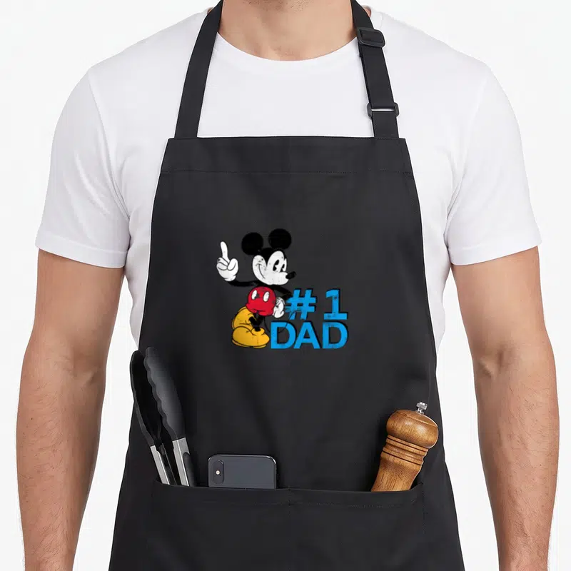 Father day apron for chefs - Mickey Mouse pointing up beside blue #1 DAD text on black background — shown on a black cotton canvas apron against a white background
