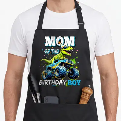 Birthday monster dinosaur truck apron - Green dinosaur in party hat behind blue monster truck with white and blue text — shown on a black cotton canvas apron against a white background