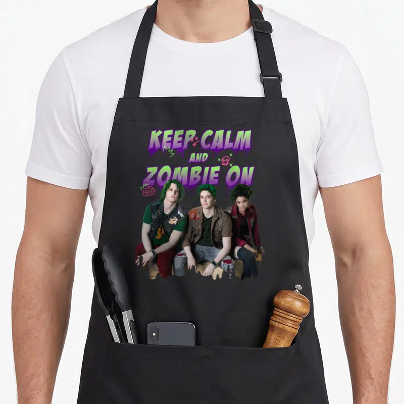 Channel zombie apron - Three green-haired zombies sit below dripping text reading KEEP CALM AND ZOMBIE — shown on a black cotton canvas apron against a white background