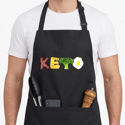 Carb keto apron - KETO text formed by bacon cheese broccoli and fried egg in red yellow green and white — shown on a black cotton canvas apron against a white background