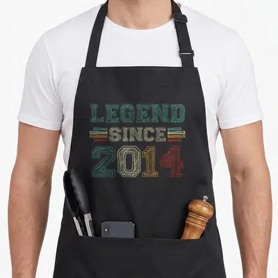 Vintage legend birthday apron - Stacked LEGEND SINCE 2014 text in teal beige red yellow distressed block letters — shown on a black cotton canvas apron against a white background
