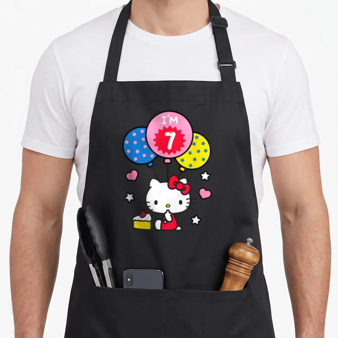 Hello general birthday apron - Hello Kitty sits with cake under three balloons, pink balloon reads I'M 7 surrounded — shown on a black cotton canvas apron against a white background
