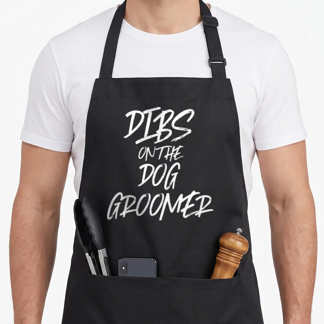 Dibss personalized dog apron - White stacked text reading Dibs On The Dog Groomer — shown on a black cotton canvas apron against a white background