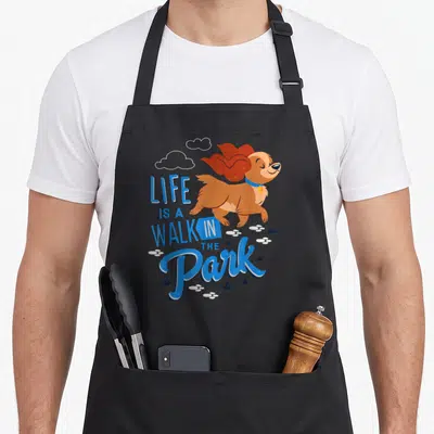 Park dog apron - Cartoon brown dog running with blue LIFE IS A WALK IN THE Park text and white paw prints — shown on a black cotton canvas apron against a white background