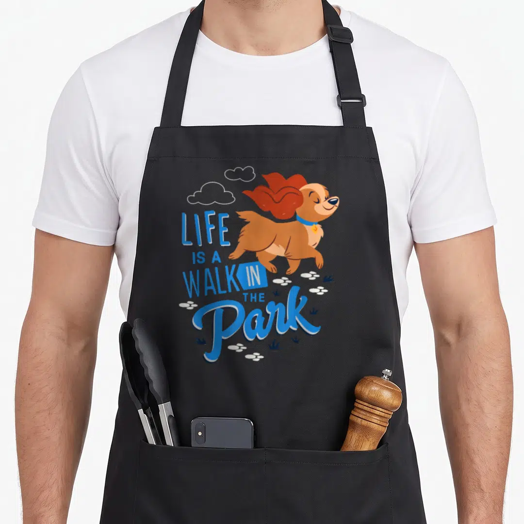 Park dog apron - Cartoon brown dog running with blue LIFE IS A WALK IN THE Park text and white paw prints — shown on a black cotton canvas apron against a white background