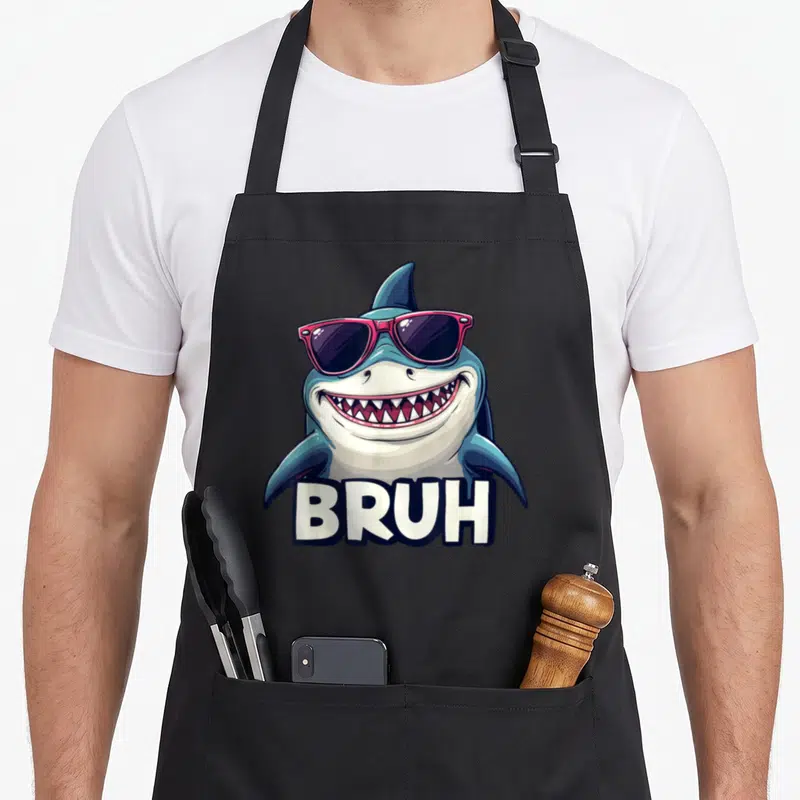 Shark streetwear apron - Cartoon shark with pink sunglasses and BRUH text in blue white pink black — shown on a black cotton canvas apron against a white background