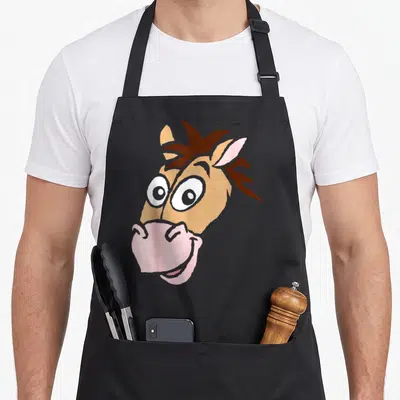 Bullseye halloween apron - Cartoon horse head in tan brown pink with large white eyes and pink muzzle centered on fabric — shown on a black cotton canvas apron against a white background
