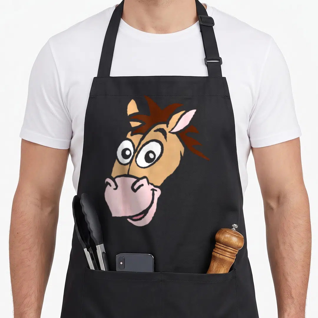 Bullseye halloween apron - Cartoon horse head in tan brown pink with large white eyes and pink muzzle centered on fabric — shown on a black cotton canvas apron against a white background