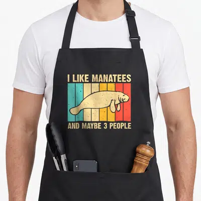 Monster cow apron - Beige manatee on blue beige yellow orange red stripes with funny text — shown on a black cotton canvas apron against a white background