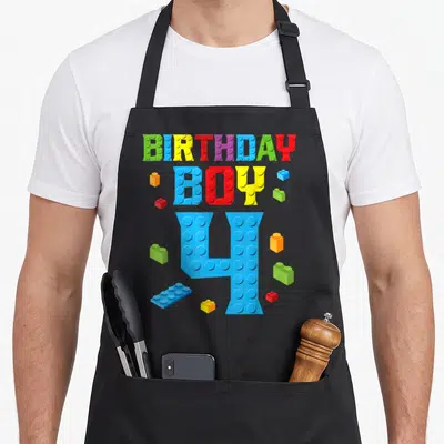 4th grill master apron - Colorful BIRTHDAY BOY and 4 text made of toy bricks with scattered blocks — shown on a black cotton canvas apron against a white background