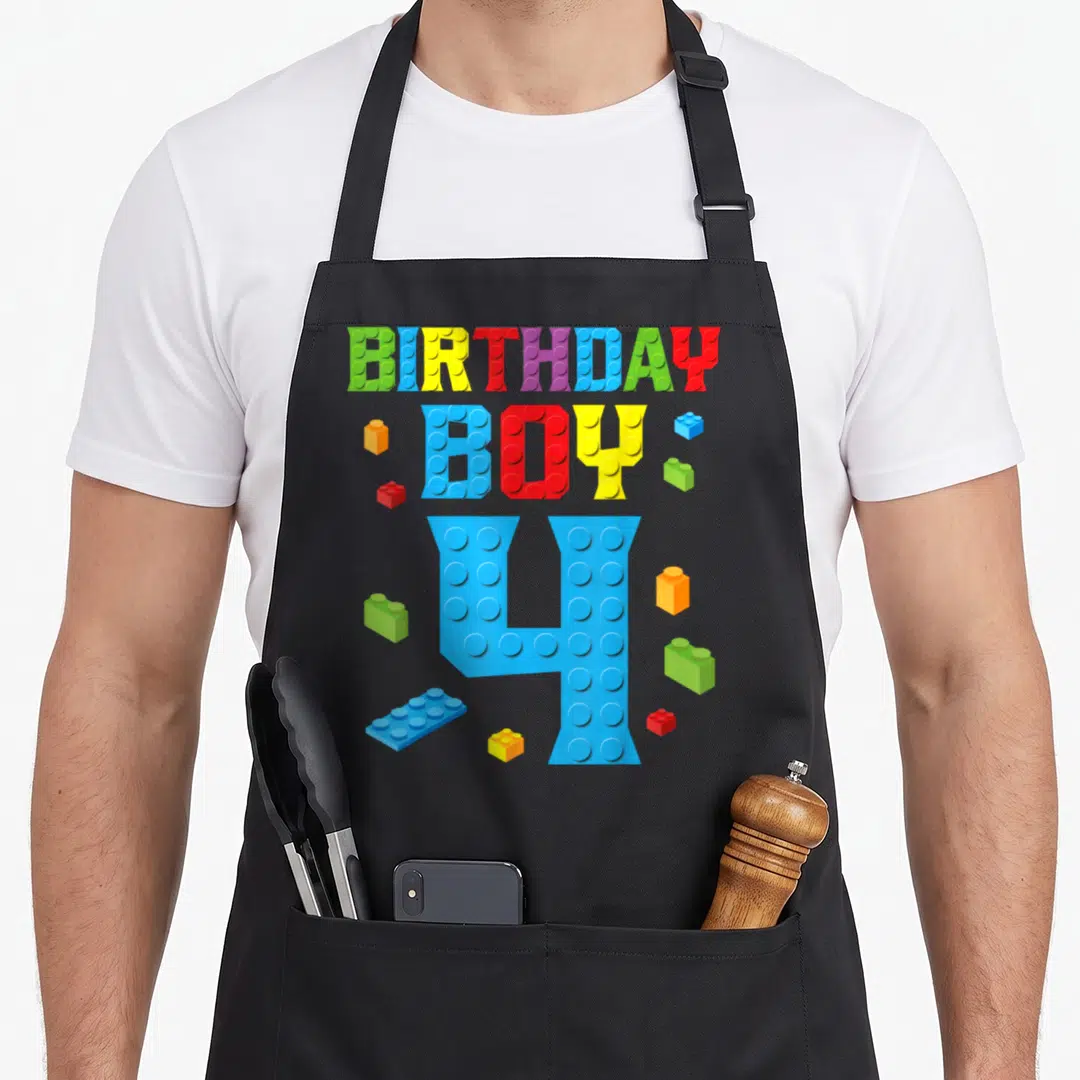 4th grill master apron - Colorful BIRTHDAY BOY and 4 text made of toy bricks with scattered blocks — shown on a black cotton canvas apron against a white background