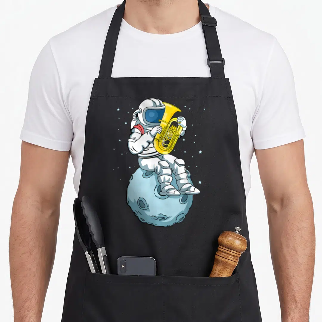 Astronaut tuba apron - Cartoon astronaut playing yellow tuba sitting on light blue moon surrounded by small stars — shown on a black cotton canvas apron against a white background