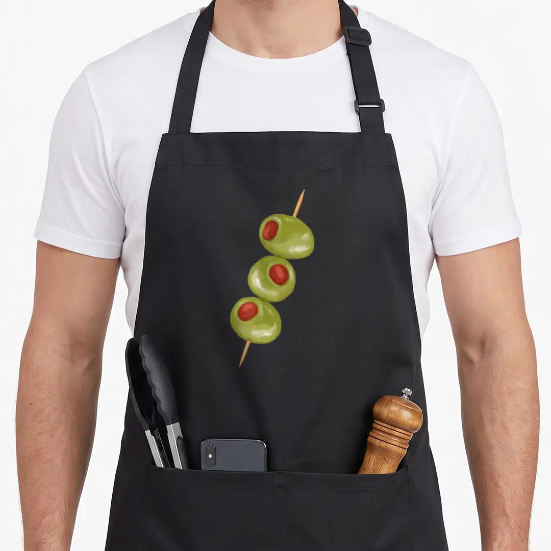 Toothpick cocktail apron - Three green olives with red centers skewered vertically on a tan wooden stick — shown on a black cotton canvas apron against a white background