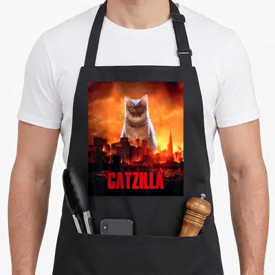 Cat kitten apron - Giant grey cat over burning city with red CATZILLA text — shown on a black cotton canvas apron against a white background