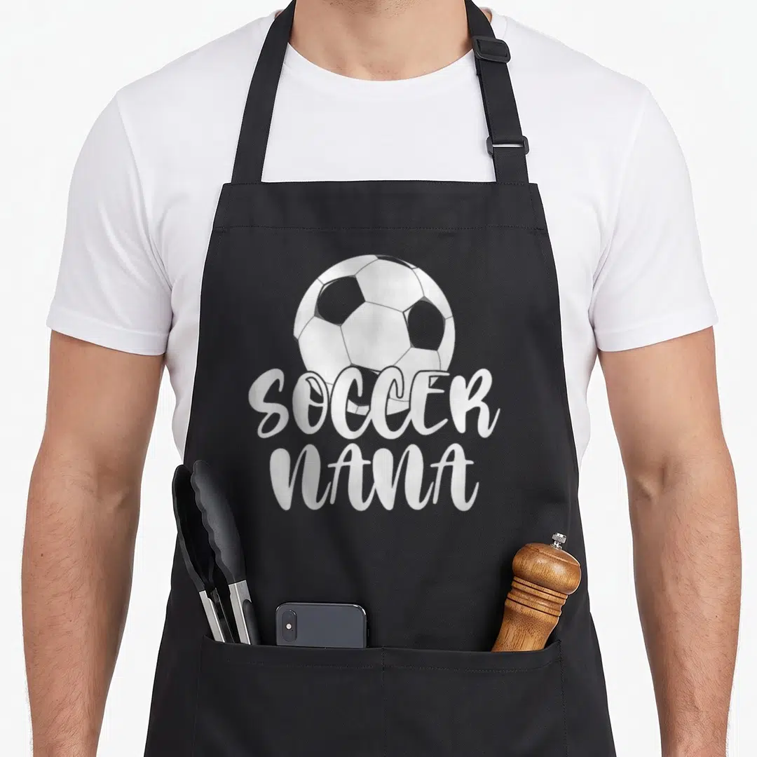 Retiree grandma aprons - White soccer ball above stacked SOCCER NANA text in cursive font — shown on a black cotton canvas apron against a white background