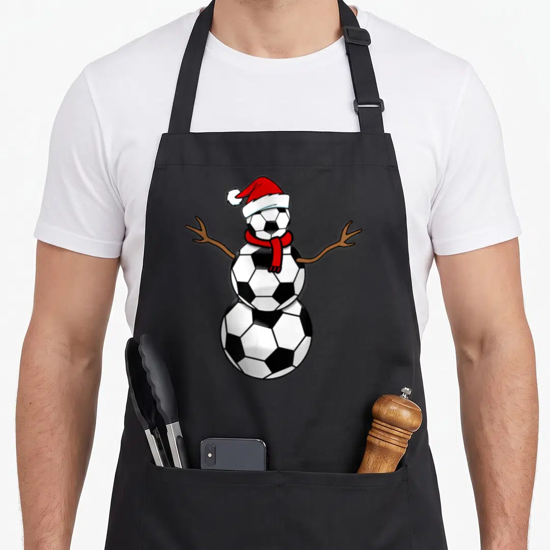 Soccer snowman apron - Cartoon snowman made of three soccer balls wearing red scarf and Santa hat — shown on a black cotton canvas apron against a white background