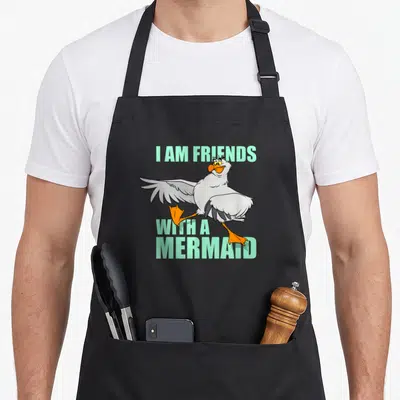 Scuttle friends apron - Cartoon seagull with orange feet and mint text I AM FRIENDS WITH A MERMAID — shown on a black cotton canvas apron against a white background