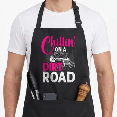 Chillin road apron - Pink and white Chillin On A Dirt Road text with UTV and pine trees — shown on a black cotton canvas apron against a white background