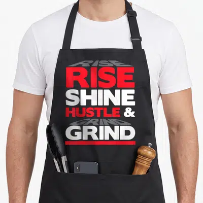 Shine rise and grind apron - Stacked red and white text reading RISE SHINE HUSTLE GRIND with red bottom bar — shown on a black cotton canvas apron against a white background