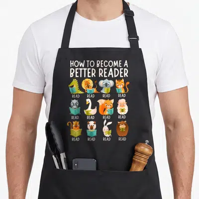 Become teacher apron - 12 cartoon animals holding books with READ text under each in 3x4 grid — shown on a black cotton canvas apron against a white background