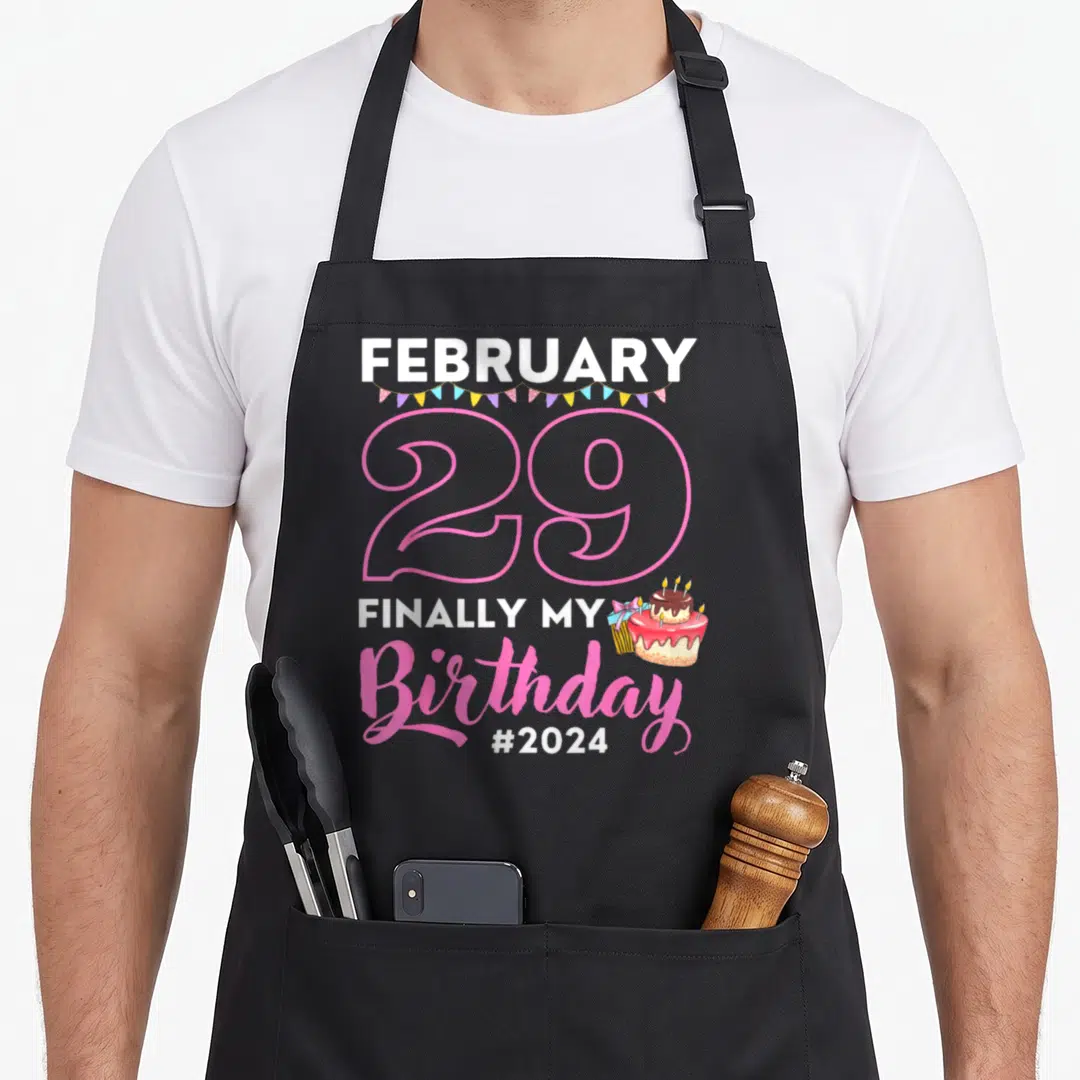 Laughter birthday apron - Stacked text FEBRUARY 29 FINALLY MY Birthday with pink outline numbers and cake graphic — shown on a black cotton canvas apron against a white background