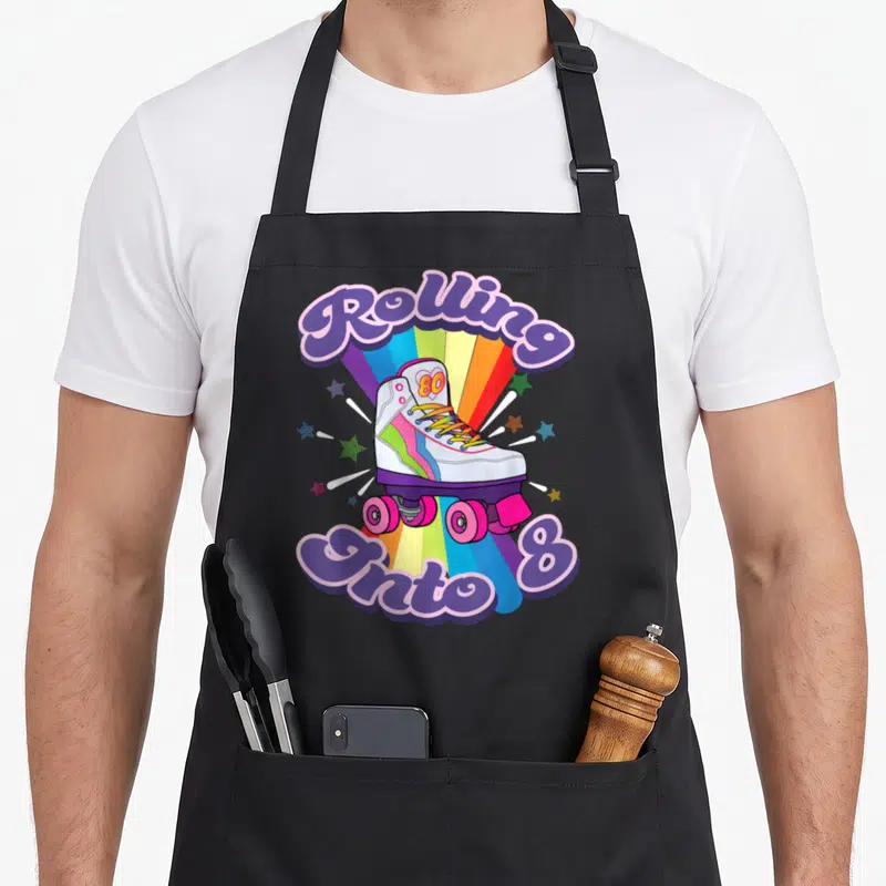 8th skate birthday apron - Cartoon roller skate on rainbow with Rolling Onto 8 text in purple and pink — shown on a black cotton canvas apron against a white background