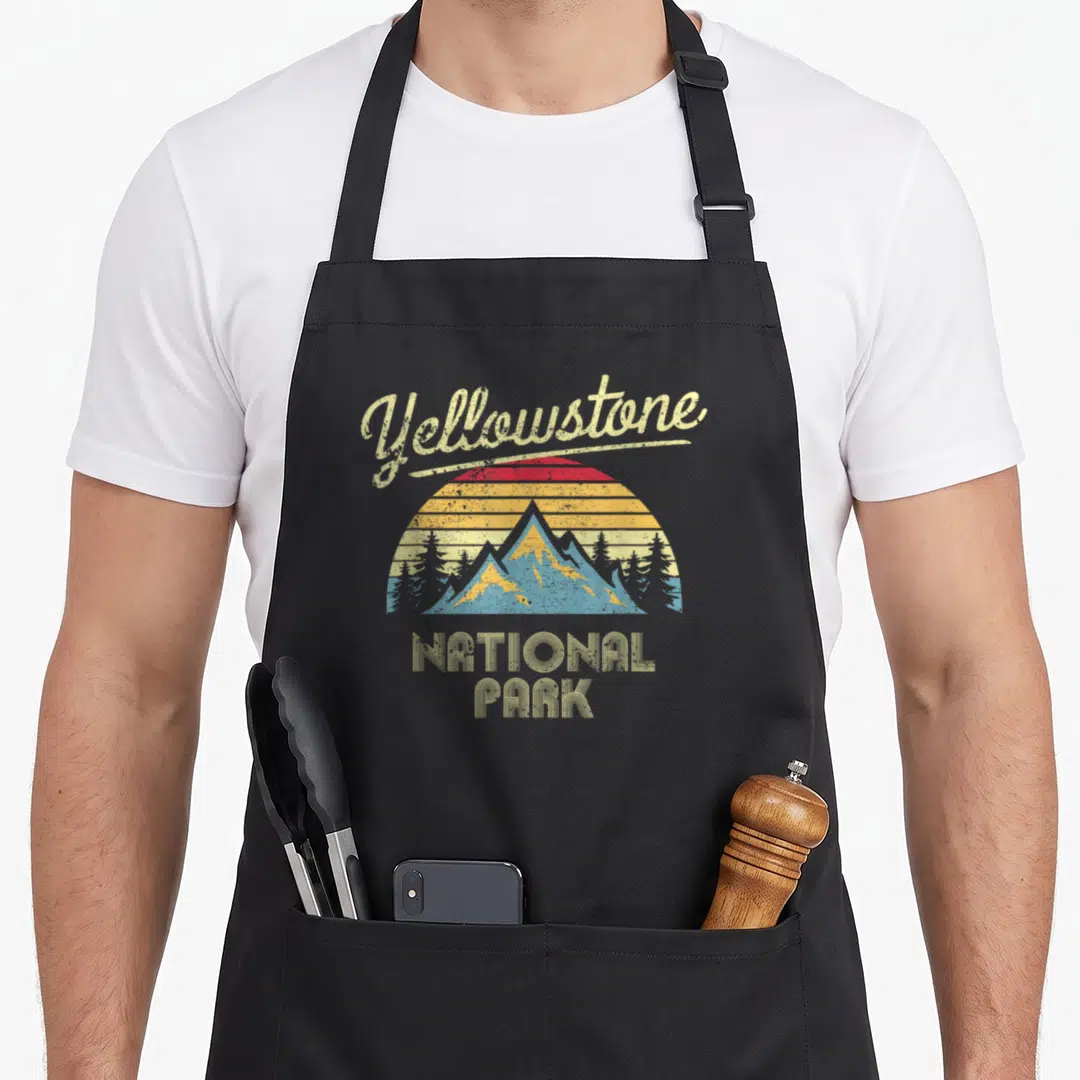 Funny national park apron - Retro Yellowstone National Park sunset mountain graphic in beige, red, orange, yellow — shown on a black cotton canvas apron against a white background