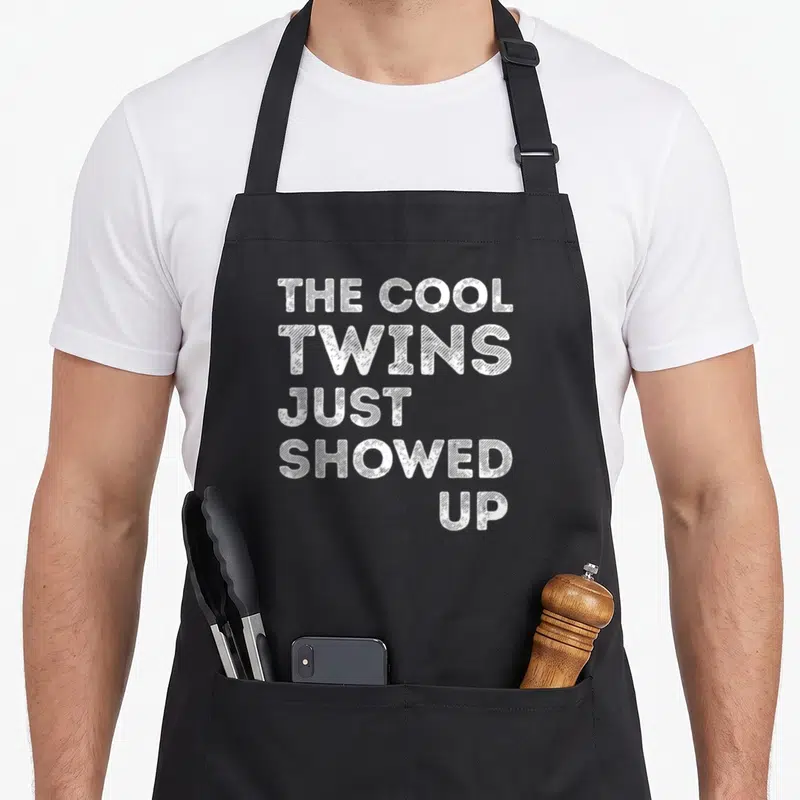 Twin boys ice apron - White stacked text reading THE COOL TWINS JUST SHOWED UP — shown on a black cotton canvas apron against a white background