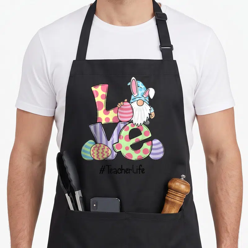 Easter teacher apron - Gnome with bunny ears behind patterned love letters and scattered eggs with hashtag text — shown on a black cotton canvas apron against a white background