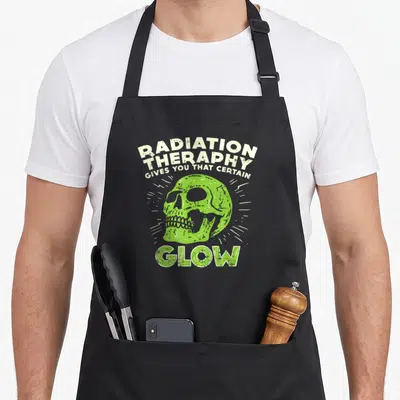 Fighter radiation aprons - Distressed green skull with white radiating lines and sarcastic text about radiation therapy — shown on a black cotton canvas apron against a white background